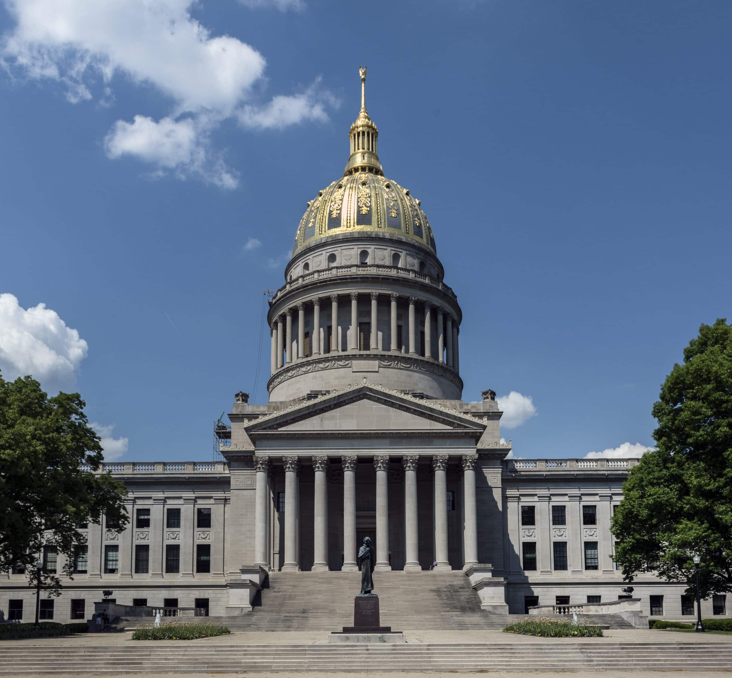 The front of the West Virginia State Capitol building is framed by trees with a statue on the stairs.